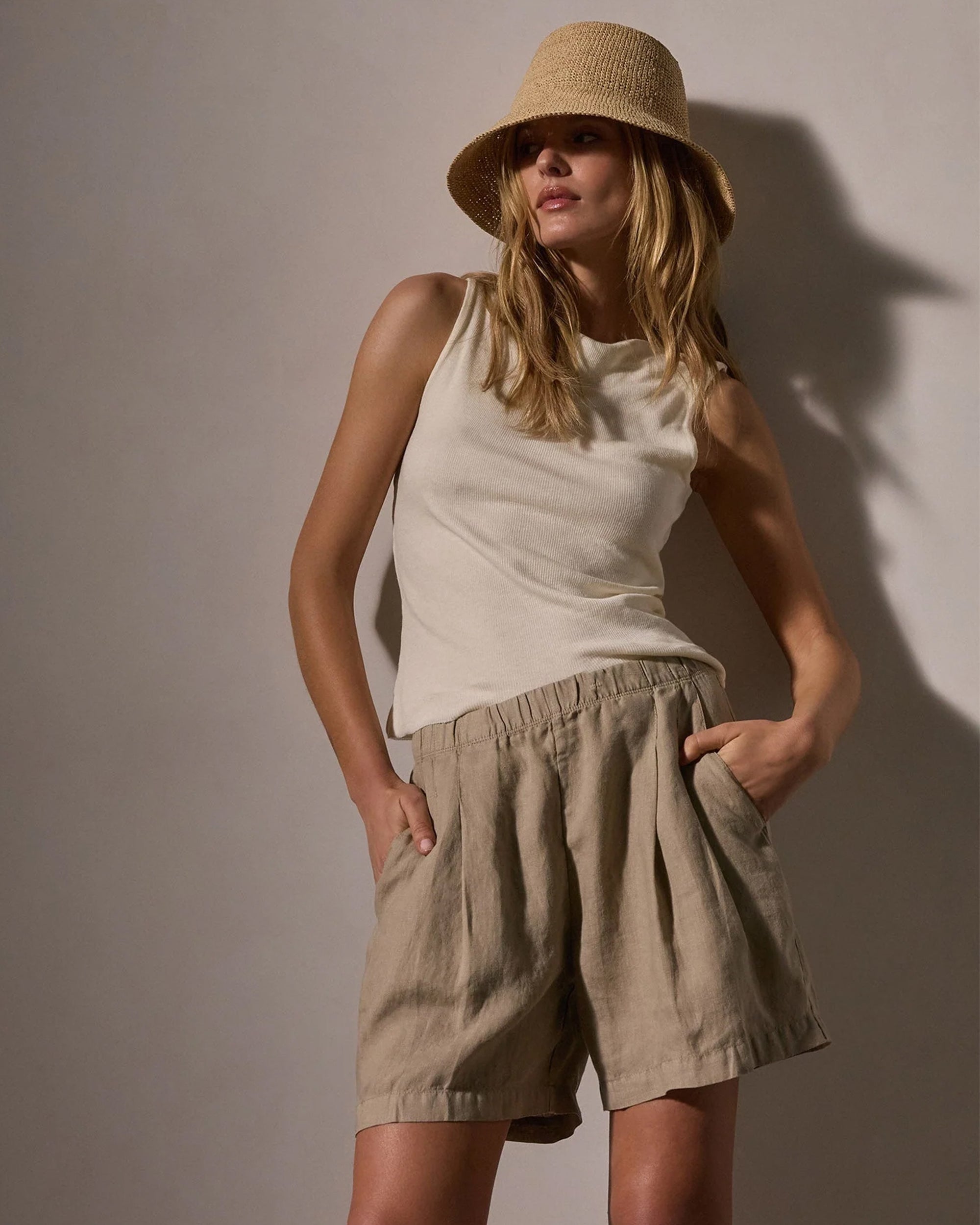 A woman stands with hands in her pockets against a neutral background, wearing James Perse Lightweight Linen Shorts, a beige bucket hat, and a sleeveless cream top.