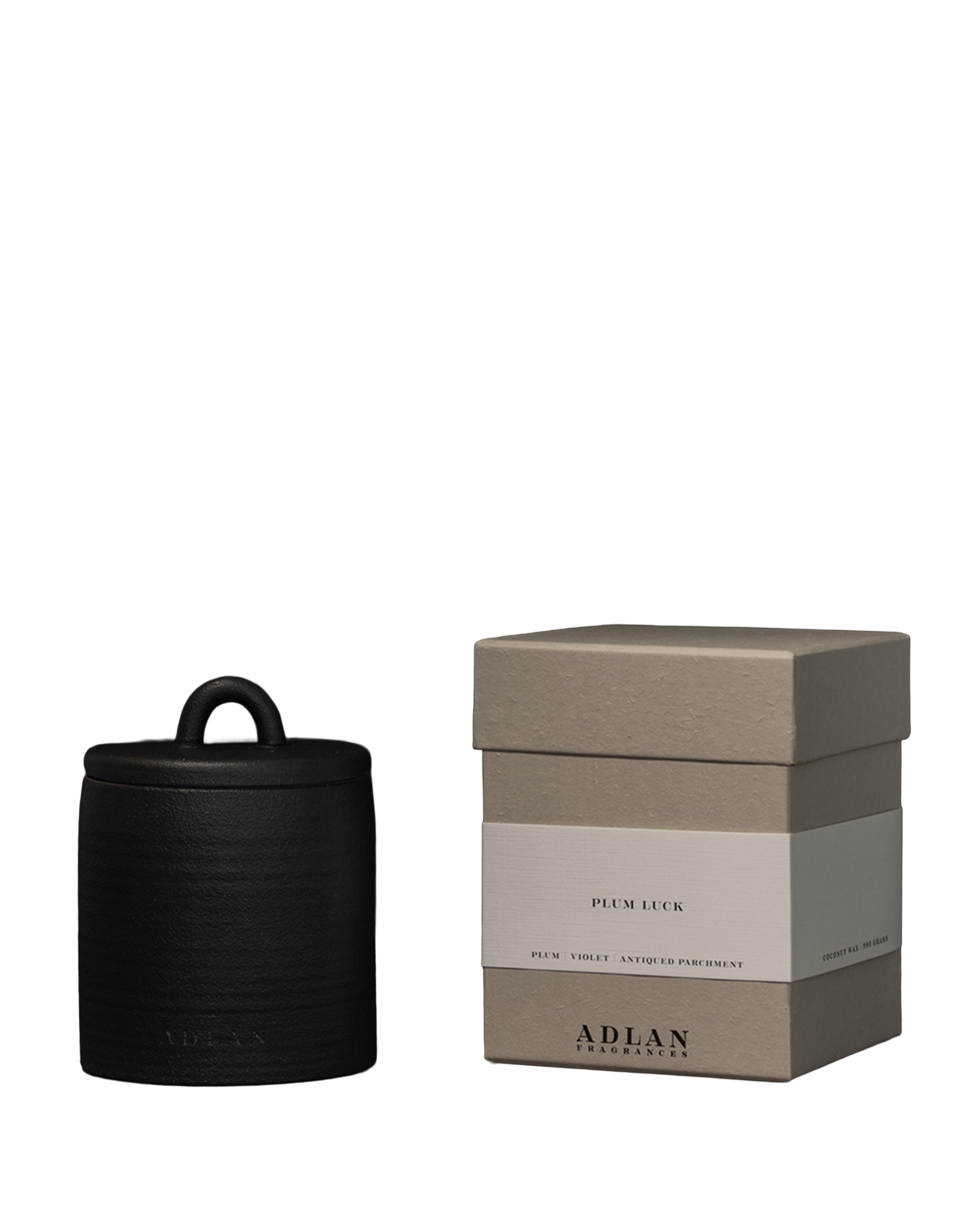 A black-lidded ADLAN Candle by ADLAN Fragrances sits beside a beige textured "Plum Luck" box, its elegant, minimalist packaging promising notes of soft violet and ripe plum inside.