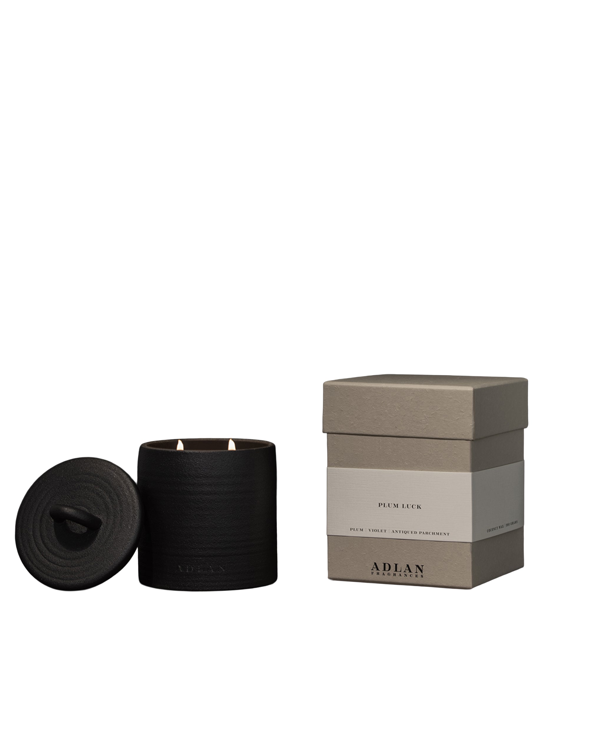 A black ADLAN Candle with a lid and two wicks sits beside a beige "ADLAN" box. The minimalist design by ADLAN Fragrances evokes luxury, blending soft violet and ripe plum notes for an elegant home scent.