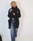 A person with long blonde hair is wearing the Poppy Zero Waste Cardigan from Kerri Rosenthal, an oversized black cotton cashmere piece with large buttons, paired with a wide scarf and distressed blue jeans. They stand against a plain white background, holding the scarf near their neck.