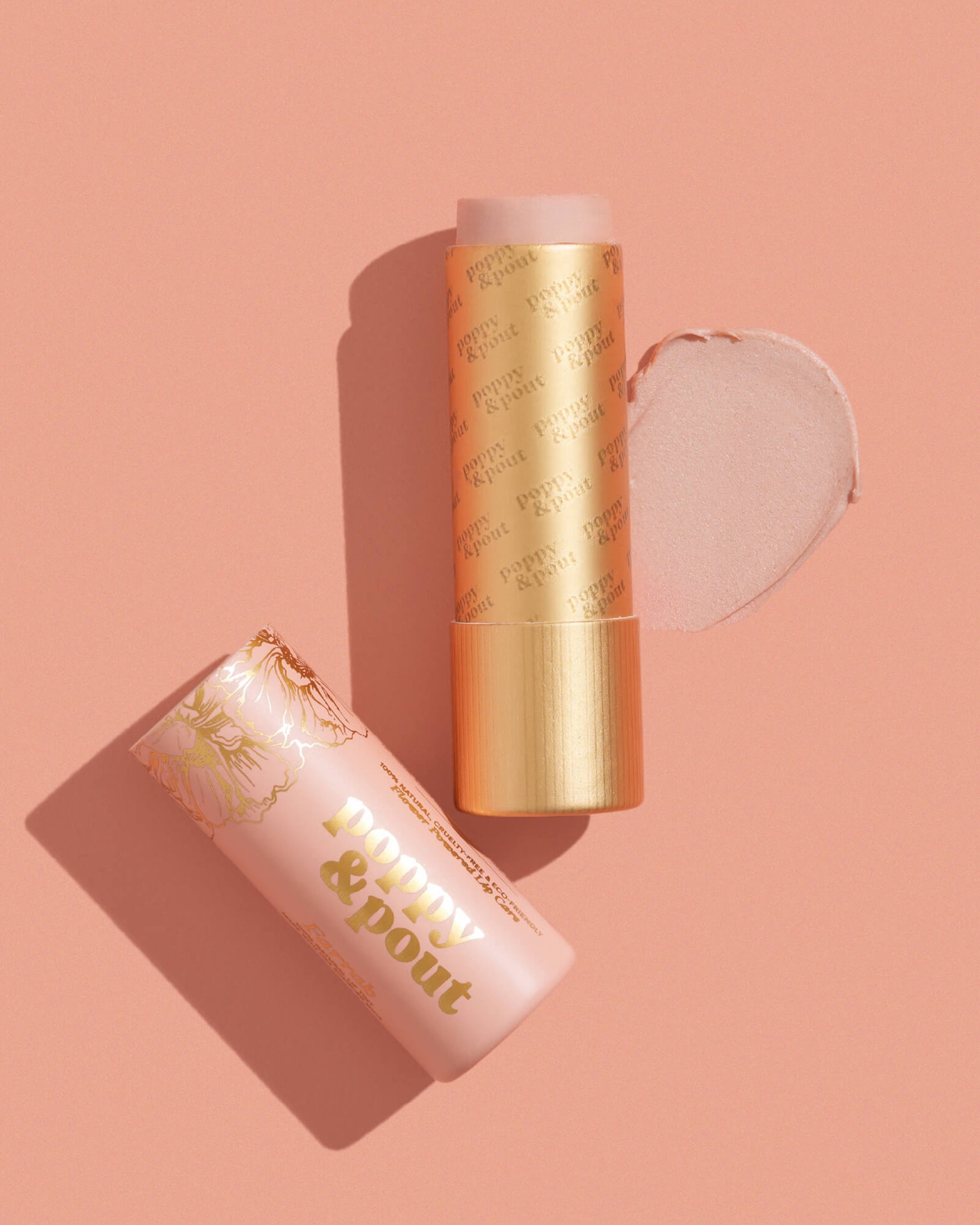 A pink and gold Poppy & Pout Lip Tint in "Farrah" by Faire lies on a peach background, its balm extended with a swipe of the pink tint shown beside it.