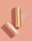 A pink and gold Poppy & Pout Lip Tint in "Farrah" by Faire lies on a peach background, its balm extended with a swipe of the pink tint shown beside it.