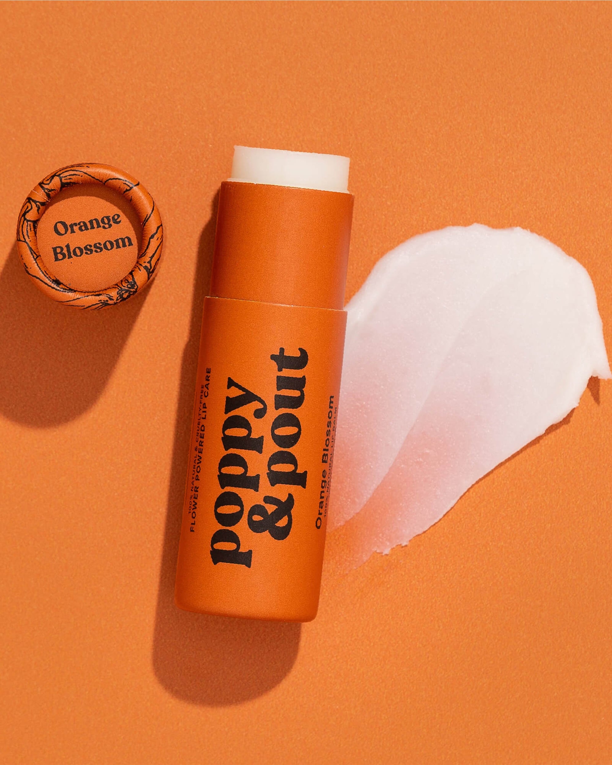 A tube of Faire Poppy & Pout Lip Balm in Orange Blossom rests on an orange surface, cap off with a swipe of balm nearby and the cap displaying the "Orange Blossom" label beside it.