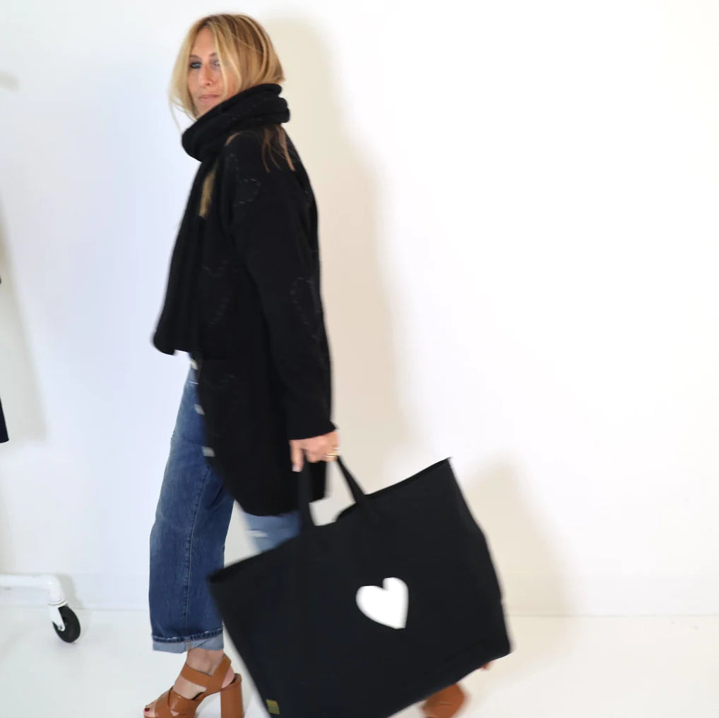 A person with long hair, dressed in a Poppy Zero Waste Cardigan by Kerri Rosenthal, a black scarf, black coat, blue jeans, and brown platform sandals, walks against a white background while carrying a black bag featuring an imperfect heart design.