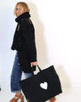 A person with long hair, dressed in a Poppy Zero Waste Cardigan by Kerri Rosenthal, a black scarf, black coat, blue jeans, and brown platform sandals, walks against a white background while carrying a black bag featuring an imperfect heart design.