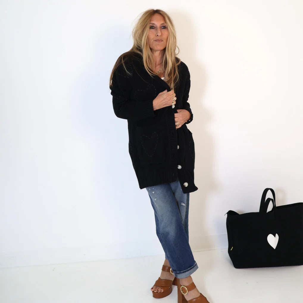 A person with long hair stands against a white background, wearing the Kerri Rosenthal Poppy Zero Waste Cardigan and blue jeans. They are barefoot beside a black bag adorned with imperfect hearts.