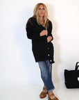 A person with long hair stands against a white background, wearing the Kerri Rosenthal Poppy Zero Waste Cardigan and blue jeans. They are barefoot beside a black bag adorned with imperfect hearts.