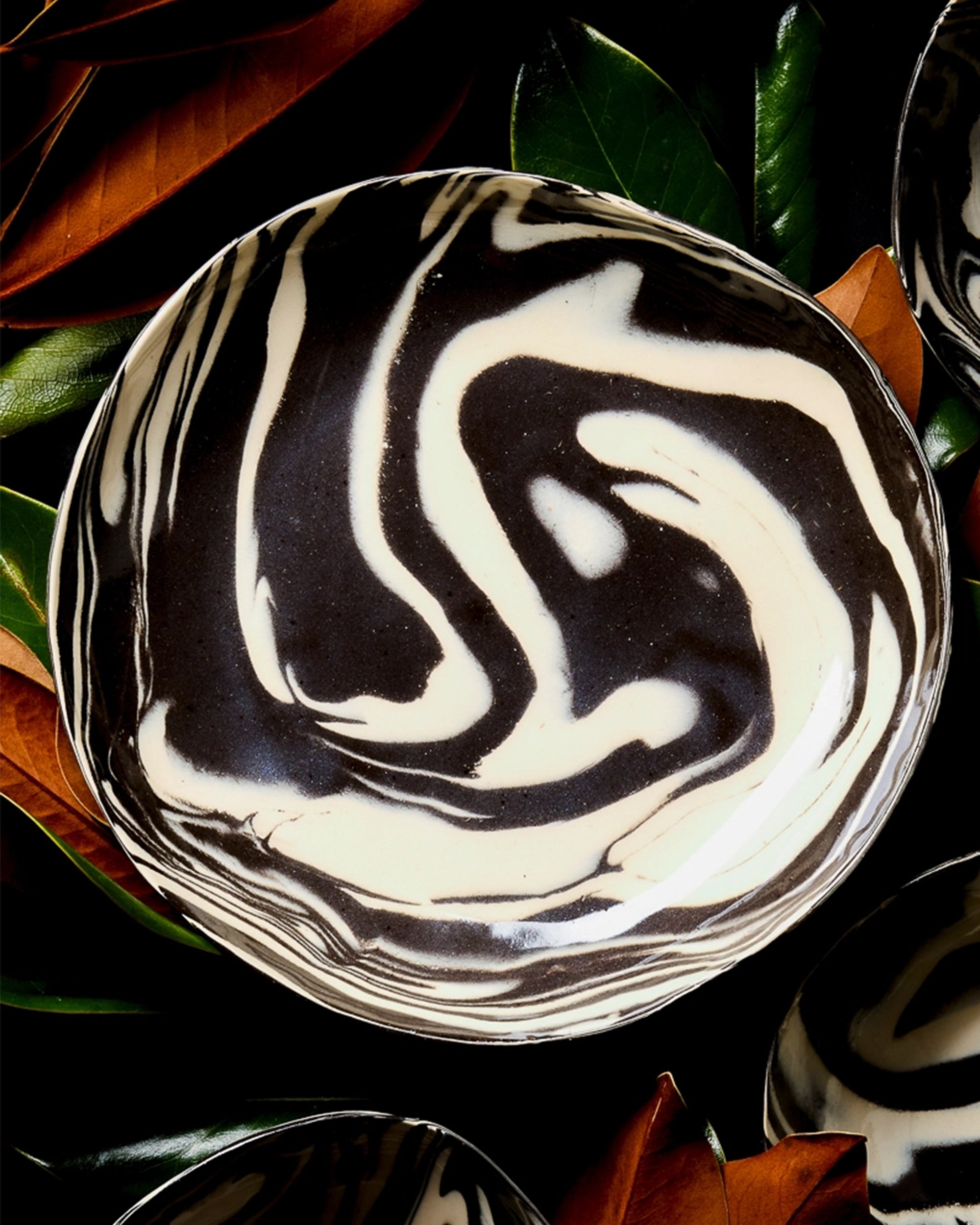 The Porcelain Serving Bowl - Black & White 10" by Faire features a bold swirling pattern, elegantly displayed atop glossy green and brown leaves.