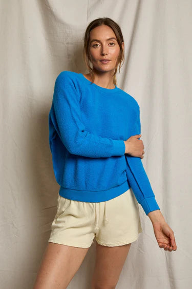 A woman stands before a beige fabric backdrop, wearing the Perfectwhitetee Ziggy Reverse LS Sweatshirt in bright blue and light drawstring shorts, one arm crossed over her body and a relaxed expression.