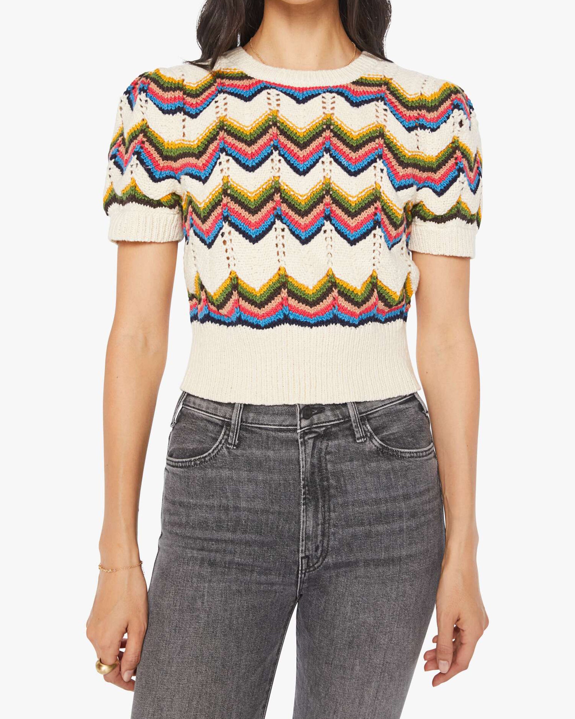 A person wears The Powder Puff knit top by Mother, featuring cream color, multicolored zigzag patterns, and puffed sleeves, styled with high-waisted black jeans. Their head is not visible.