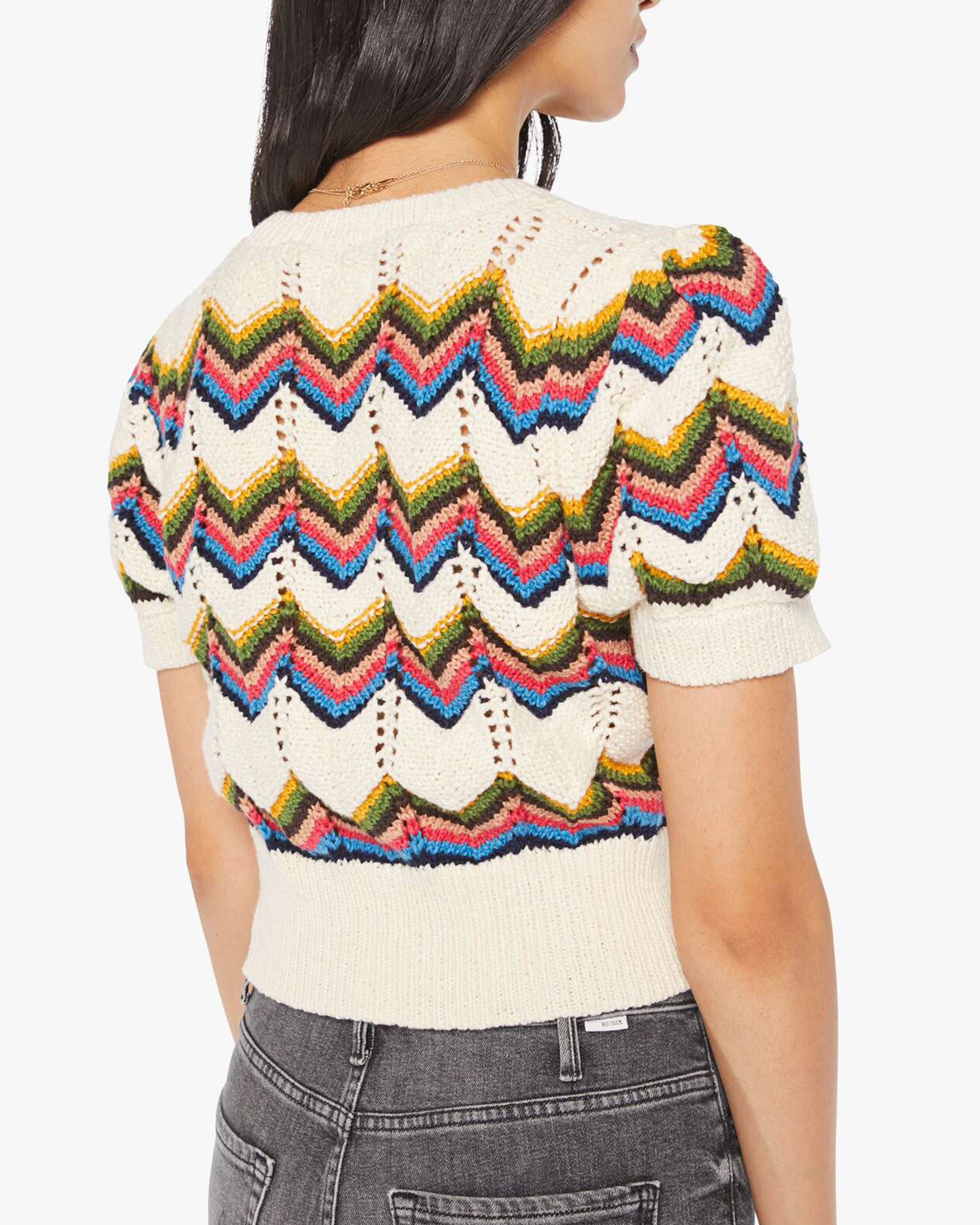 A woman with long dark hair is shown from the back wearing The Powder Puff by Mother, a cropped short-sleeve knit top with colorful zigzag patterns, paired with high-waisted gray jeans.
