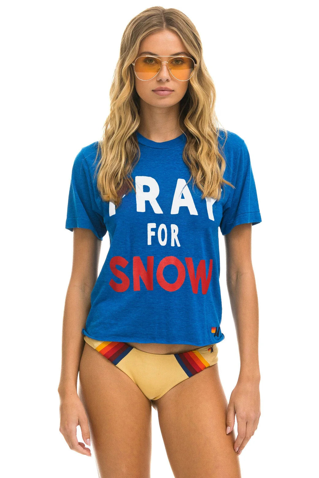 A woman with long blonde hair sports vintage-style orange sunglasses and a blue Aviator Nation "Pray for Snow" boyfriend tee. She pairs it with yellow bikini bottoms adorned with red and black stripes, standing against a white background.