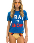 A woman with long blonde hair sports vintage-style orange sunglasses and a blue Aviator Nation "Pray for Snow" boyfriend tee. She pairs it with yellow bikini bottoms adorned with red and black stripes, standing against a white background.