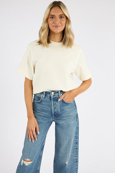 A woman with blonde hair smiles with one hand in her pocket, wearing the Citizens Of Humanity/AGOLDE Goldie Tee—an oversized, ribbed cotton blend cream t-shirt—with high-waisted blue jeans featuring ripped knees, against a plain white background.