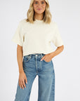 A woman with blonde hair smiles with one hand in her pocket, wearing the Citizens Of Humanity/AGOLDE Goldie Tee—an oversized, ribbed cotton blend cream t-shirt—with high-waisted blue jeans featuring ripped knees, against a plain white background.