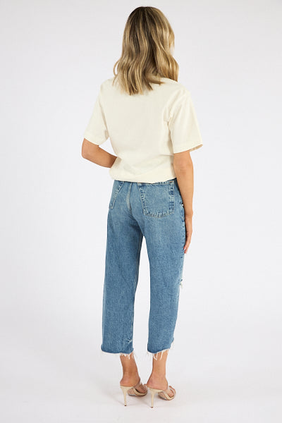 A woman with shoulder-length blonde hair faces away, wearing the Citizens Of Humanity/AGOLDE Goldie Tee in beige, high-waisted blue jeans with raw hems, and beige heeled sandals against a plain white background.