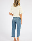 A woman with shoulder-length blonde hair faces away, wearing the Citizens Of Humanity/AGOLDE Goldie Tee in beige, high-waisted blue jeans with raw hems, and beige heeled sandals against a plain white background.