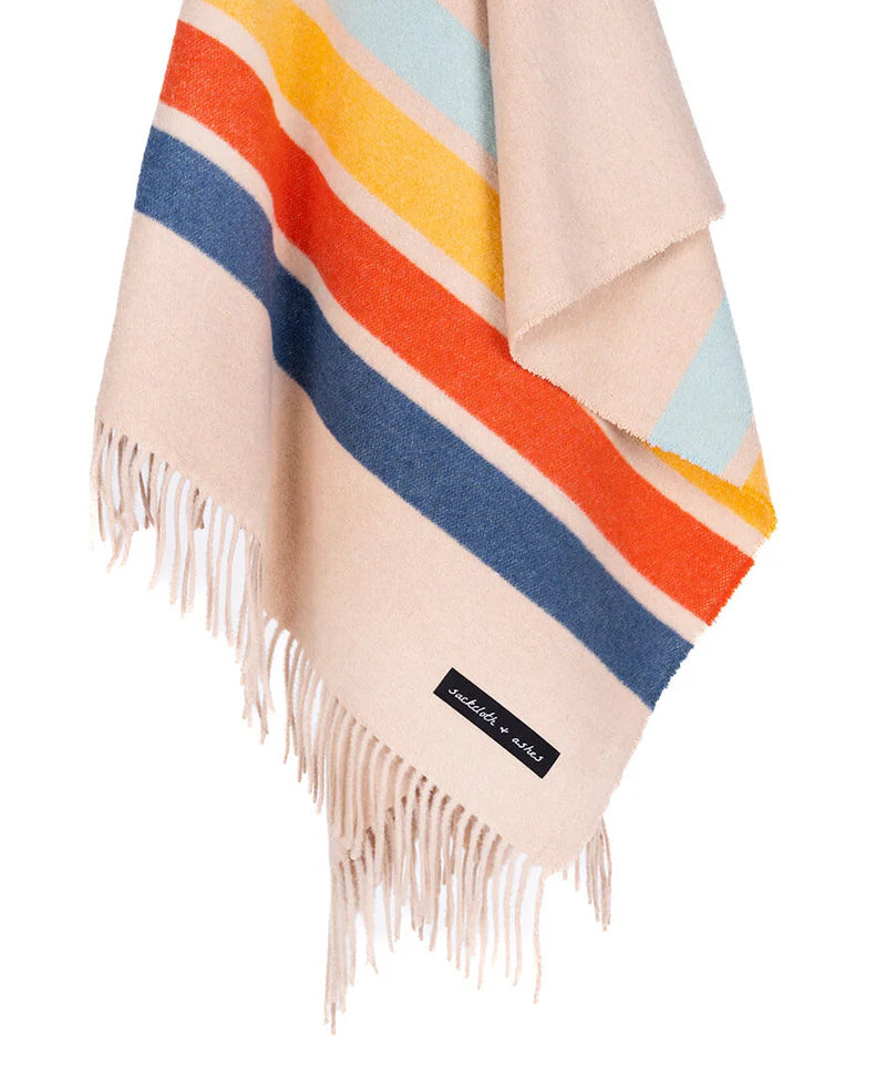 The Sackcloth & Ashes/Camp Coast by Sackcloth & Ashes is a beige wool blanket made from recycled materials, with fringed edges, horizontal stripes in blue, orange, red, and light blue, and a small black label near the bottom.