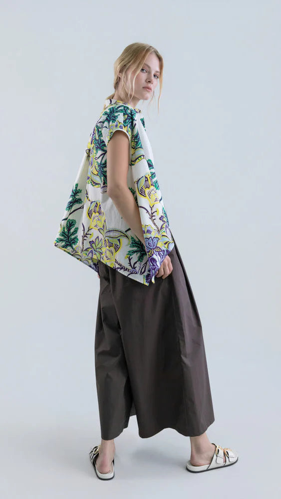 A woman with blonde hair tied back wears a loose floral cotton poplin top, Psophia’s Air Drift Wide-Leg Culotte with Drawstring Waist in dark brown, and white slip-on shoes as she poses sideways against a plain light background.