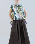 A woman wears the Psophia Air Drift Wide-Leg Culotte with Drawstring Waist, a loose, dark brown cotton poplin culotte, paired with a colorful floral sleeveless top and white slide sandals, standing against a plain light background.
