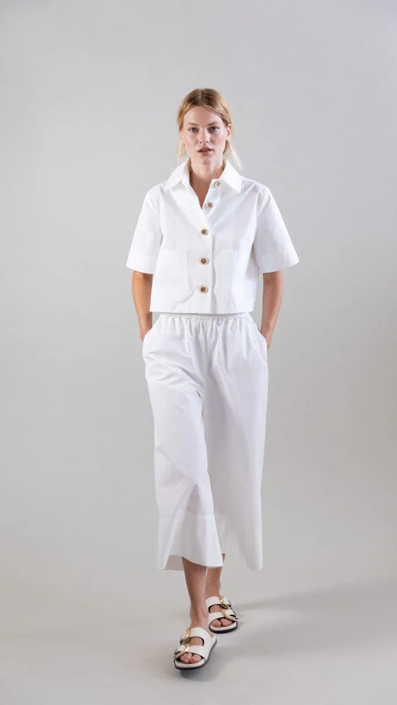 A person stands against a plain background wearing a white short-sleeved button-up shirt, Psophia's Bouton & Brume Elastic-Waist Cotton Palazzo Pants, and white sandals with gold accents, hands in their pockets.