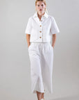A person stands against a plain background wearing a white short-sleeved button-up shirt, Psophia's Bouton & Brume Elastic-Waist Cotton Palazzo Pants, and white sandals with gold accents, hands in their pockets.