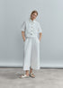 A woman poses against a gray backdrop, wearing the Psophia 2716 Womens Trouser—cotton palazzo pants with an elastic waistband—and a short-sleeved white button-up shirt, finishing the look with white slide sandals and hands in her pockets.