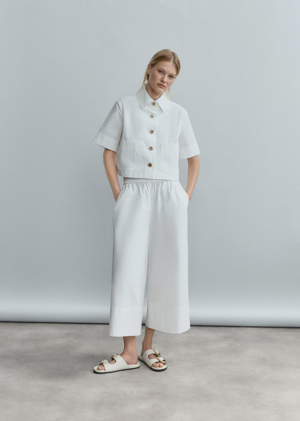 A woman stands against a light grey backdrop wearing Psophia’s Bouton & Brume Elastic-Waist Cotton Palazzo Pants, paired with a white short-sleeve button-up shirt and white slide sandals with decorative buckles, hands in her pockets.