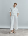 A woman stands against a light grey backdrop wearing Psophia’s Bouton & Brume Elastic-Waist Cotton Palazzo Pants, paired with a white short-sleeve button-up shirt and white slide sandals with decorative buckles, hands in her pockets.