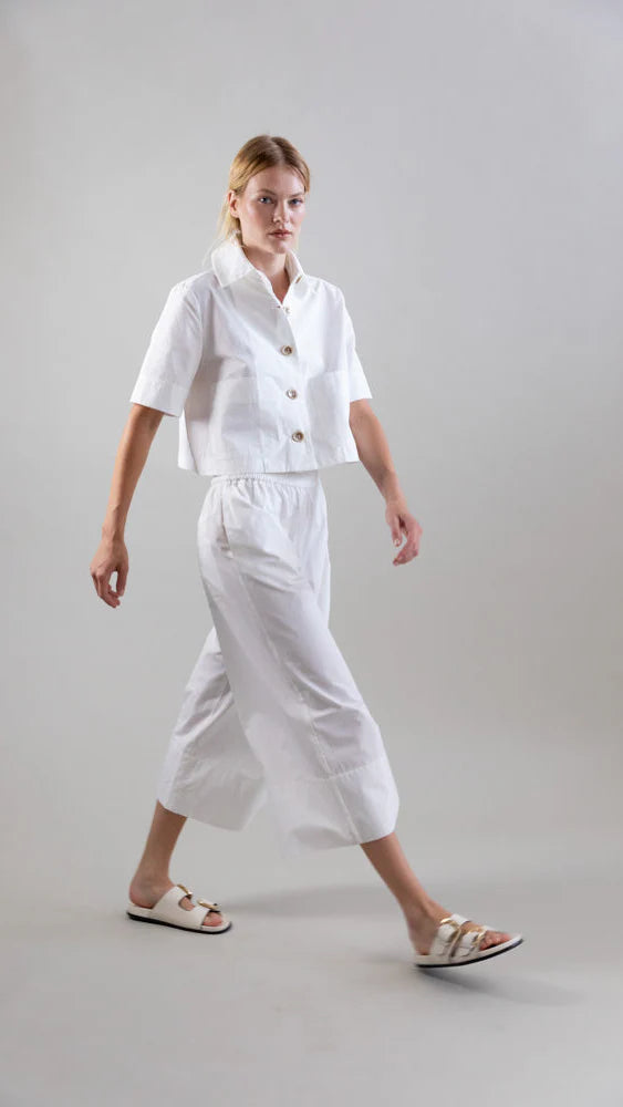 A person in a white short-sleeve button-up shirt, Psophia’s Bouton & Brume Elastic-Waist Cotton Palazzo Pants, and white sandals walks confidently against a gray background, showcasing the pants' flowing silhouette.