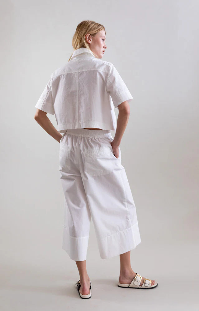 A person faces away, wearing Psophia's Bouton & Brume Short-Sleeve Cotton Bolero over a loose white shirt, with matching wide-leg cropped pants and white slide sandals against a plain light background.