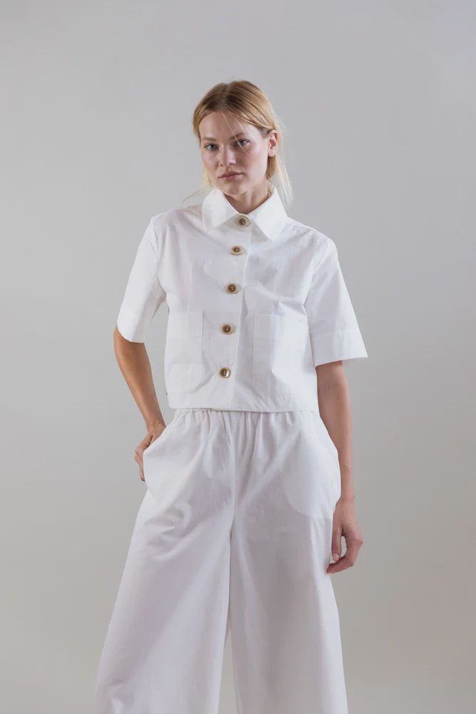 A blonde woman stands against a plain backdrop, wearing the Psophia Bouton & Brume Short-Sleeve Cotton Bolero with matching wide-leg pants. She looks directly at the camera, hands in her pockets.