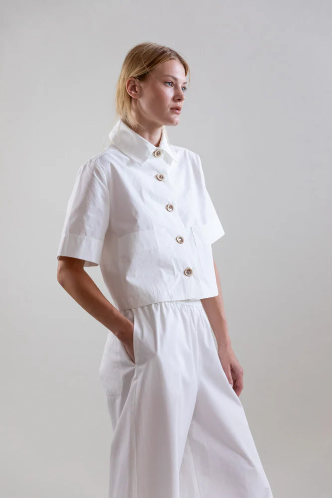 A woman stands in a white studio wearing the Psophia Bouton & Brume Short-Sleeve Cotton Bolero over a collared white shirt and loose white pants, her hands in her pockets as she gazes thoughtfully to the side.