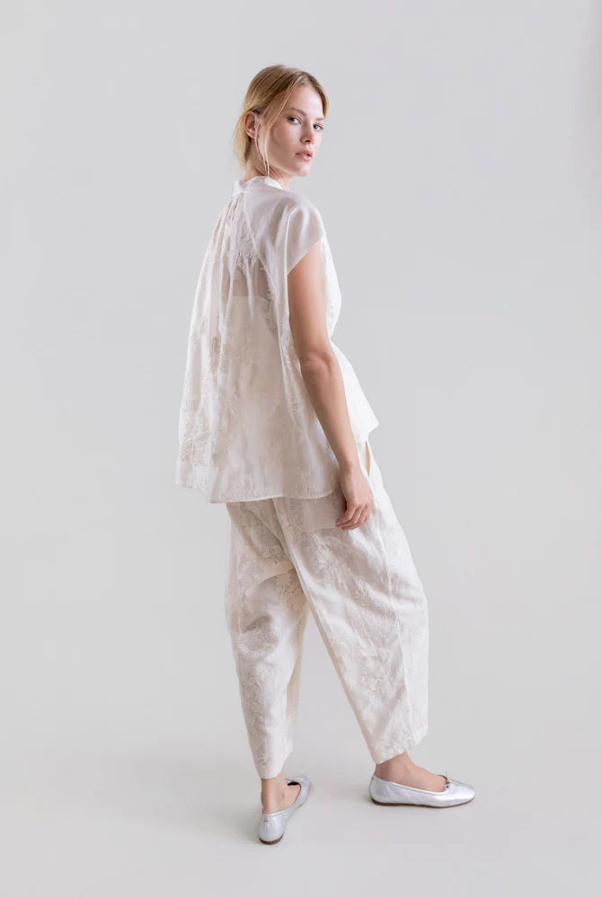 A woman stands against a plain light background, looking over her shoulder. She wears the Psophia Ethereal Botanical Tencel Ring-Neck Blouse with short sleeves, matching white textured pants, and white ballet flats.
