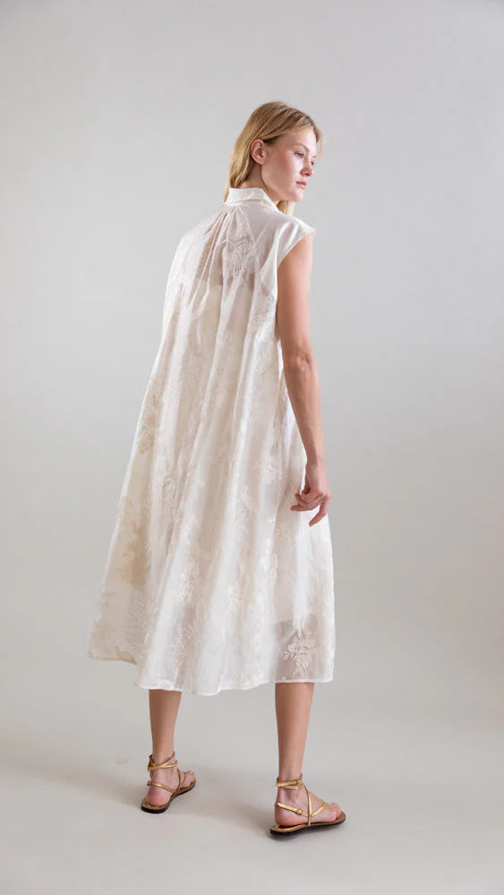 A woman stands on a light background, wearing the Psophia Ethereal Botanical Tencel Shirt-Inspired Dress—a white, sleeveless, knee-length design with delicate floral embroidery—and gold flat sandals. She is turned slightly, looking over her shoulder.