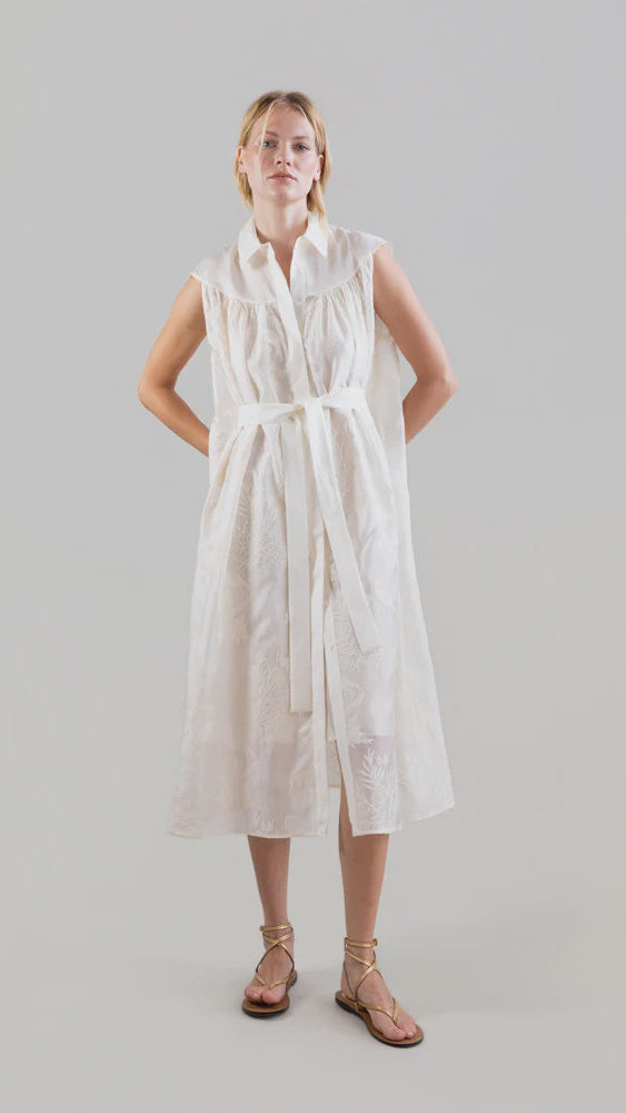 A person stands against a plain background, wearing the Psophia Ethereal Botanical Tencel Shirt-Inspired Dress—sleeveless, white, knee-length with embroidery, belted waist, collar, and subtle texture. They're also in flat strappy sandals with hands behind their back.