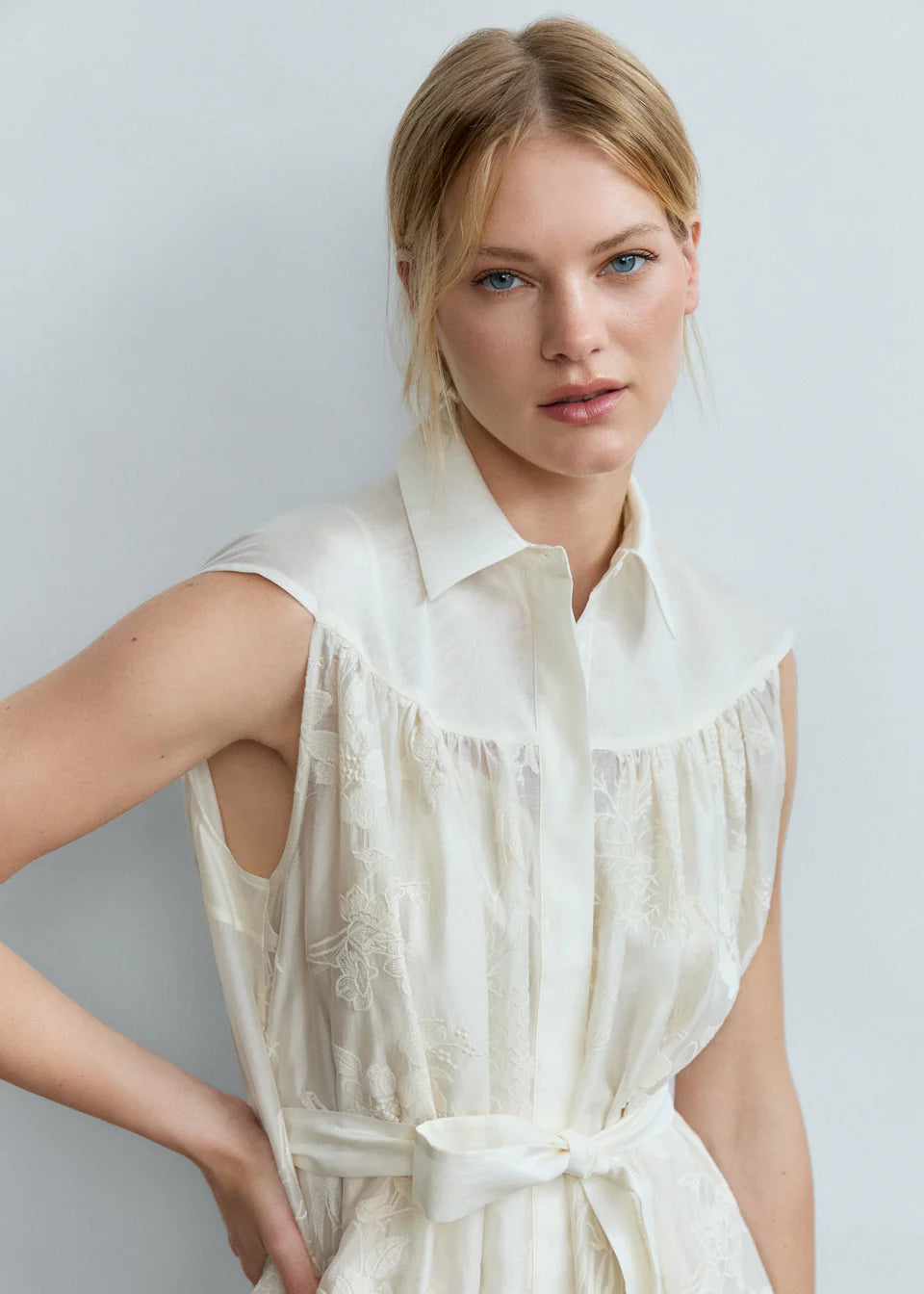 A woman with blond hair in a low ponytail wears the Psophia Ethereal Botanical Tencel Shirt-Inspired Dress, featuring floral embroidery, a belted waist, and an A-line silhouette, standing against a light gray background.