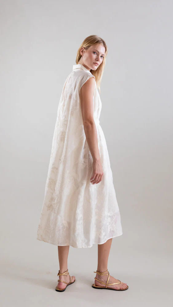 A woman in the Psophia Ethereal Botanical Tencel Shirt-Inspired Dress, featuring a sleeveless A-line silhouette and high collar, stands barefoot on light flooring, glancing over her shoulder against a plain backdrop.
