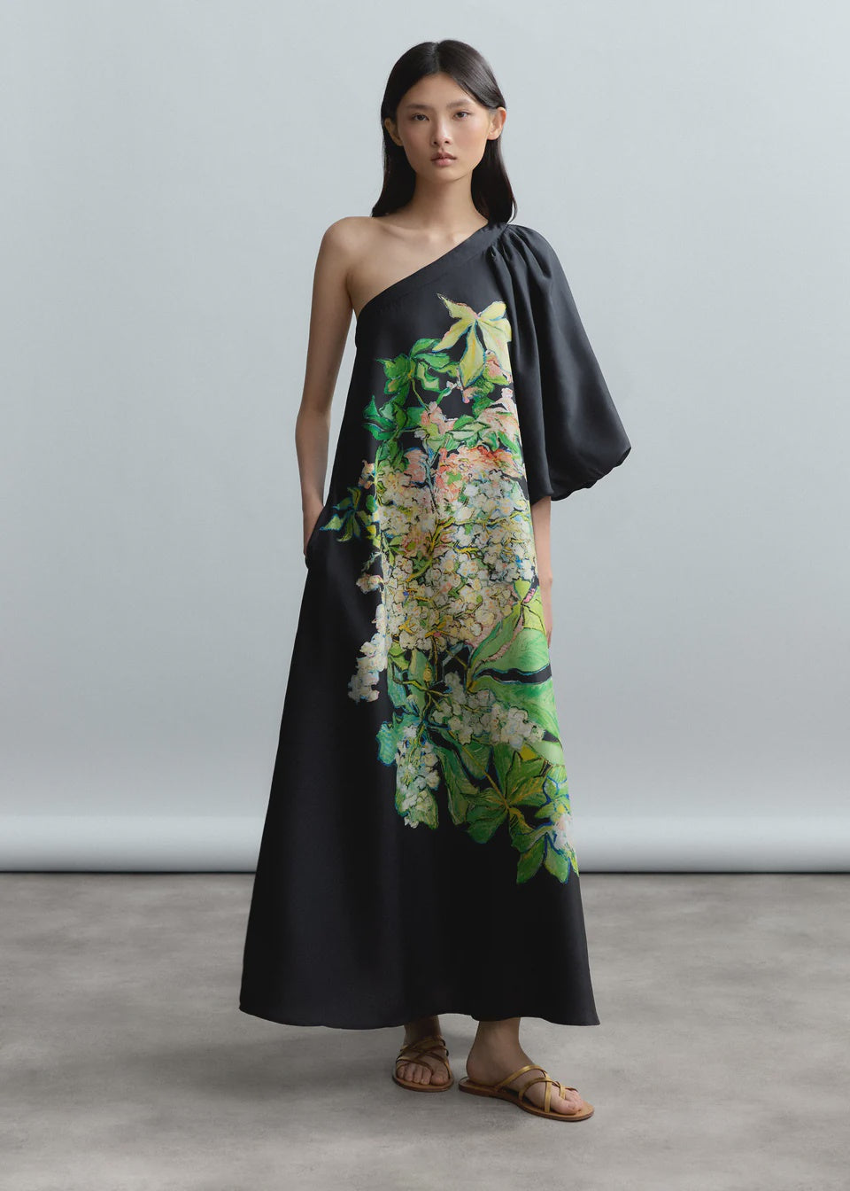 A woman wears the Psophia One Shoulder Dress in silk habotai with an asymmetrical neckline, single loose sleeve, and Hydrangea Brush print in green, yellow, and white, styled with simple sandals against a neutral background.