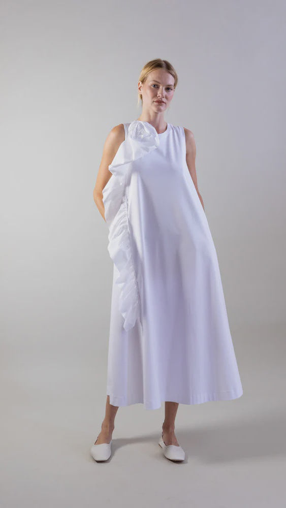 A woman wears the Psophia Sculptural Rosette Mercerized Cotton Midi Dress in white, featuring a side rosette detail, paired with white flat shoes, standing against a plain light gray background.