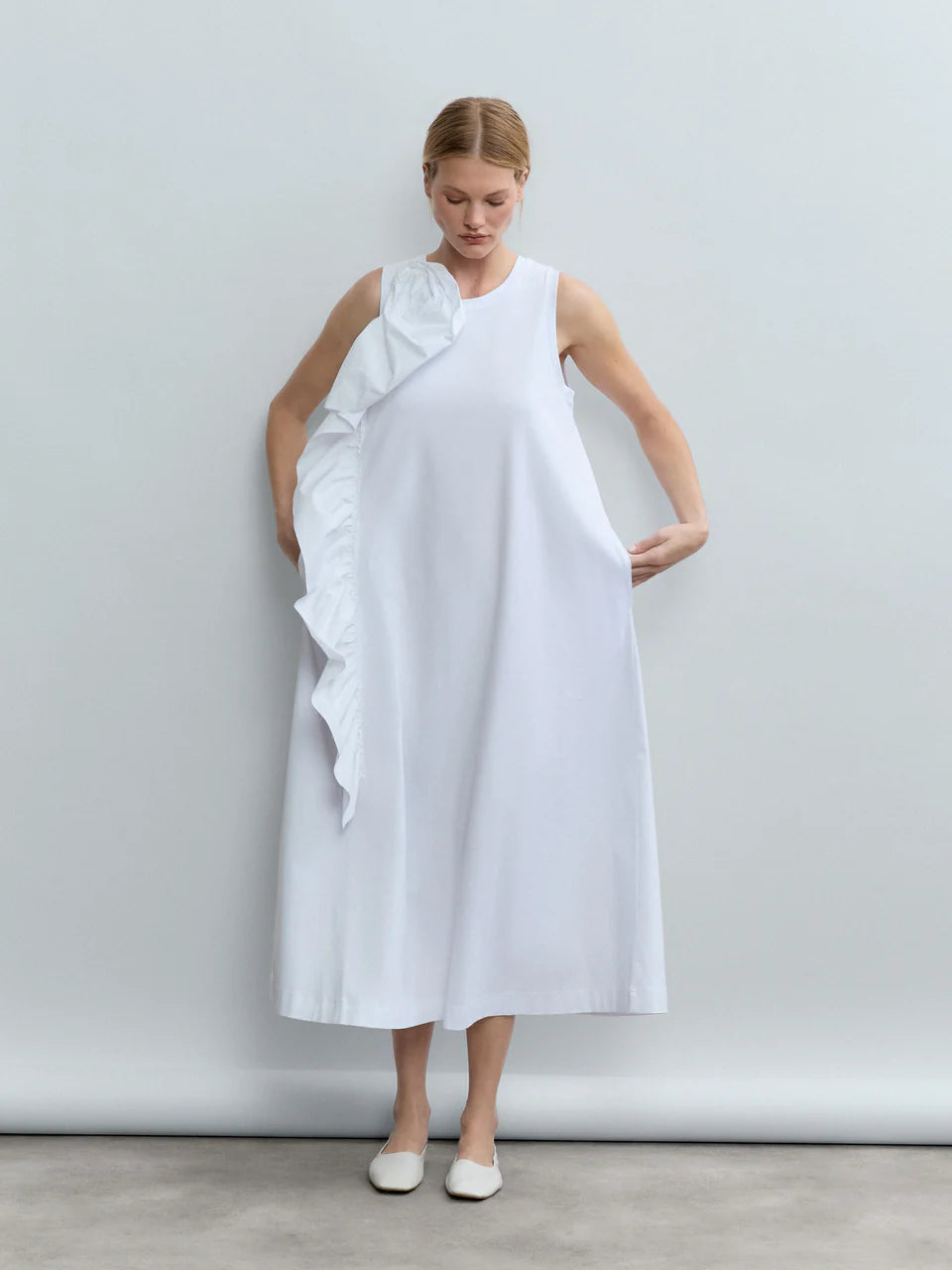 A woman in the Psophia Sculptural Rosette Mercerized Cotton Midi Dress stands against a plain gray background, looking down and gently holding the sides. The sleeveless dress features a ruffled sculptural rosette along the left shoulder and side.