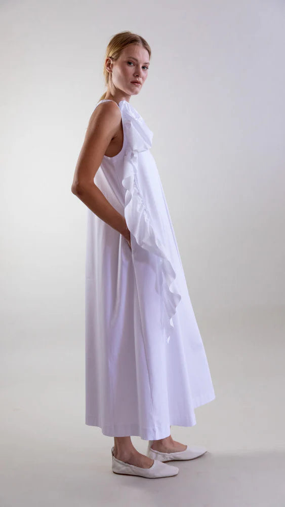 A woman stands in profile, hands in pockets, wearing the Psophia Sculptural Rosette Mercerized Cotton Midi Dress with front ruffled detailing and white flats against a simple light background.