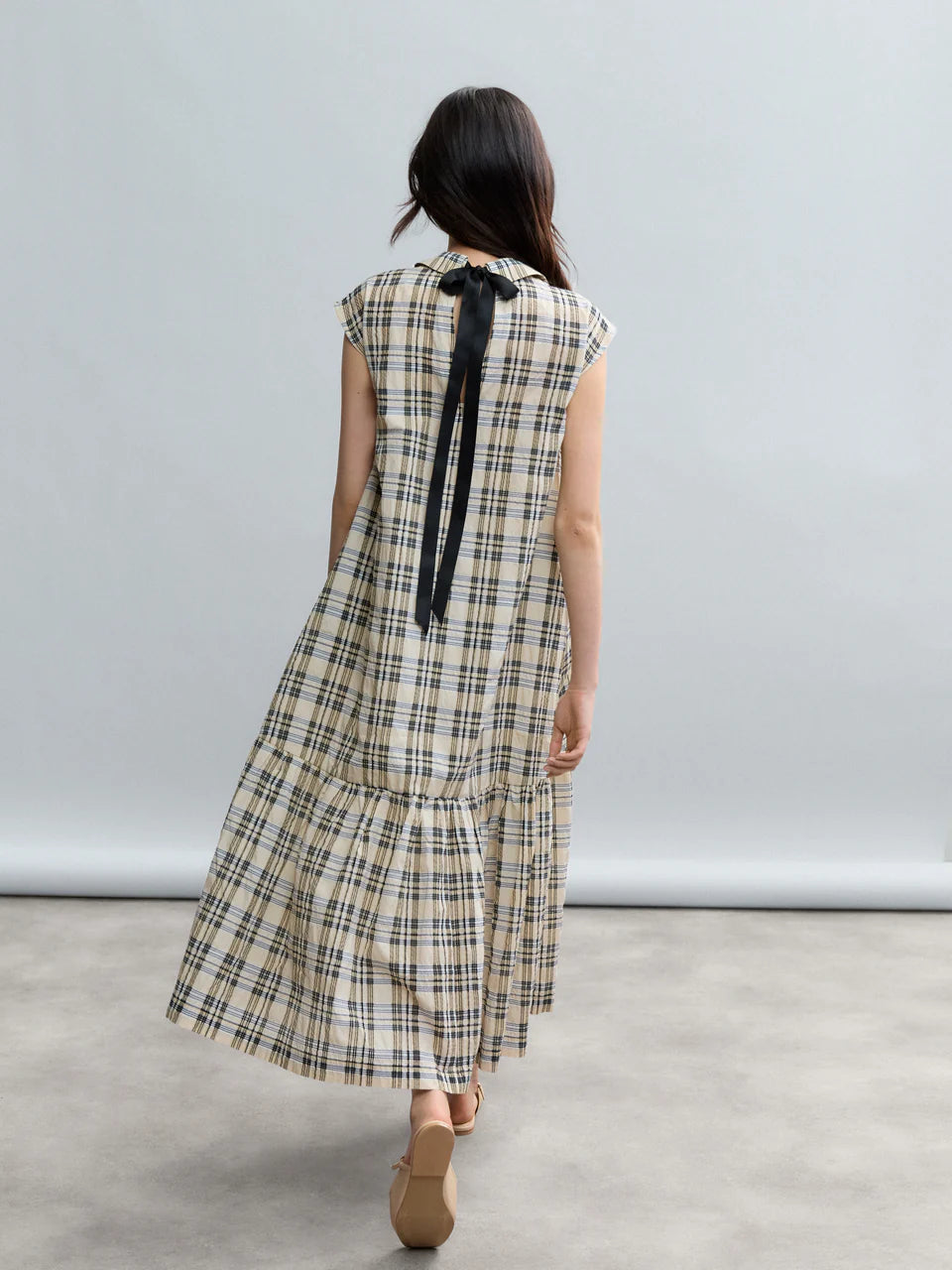 A woman with long dark hair stands on a gray floor, facing away, wearing the Psophia Soft-Touch Plaid A-Line Drop-Waist Dress against a light gray background.