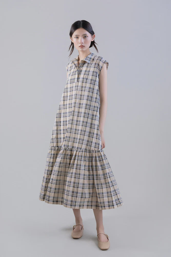 A young woman wears the Psophia Soft-Touch Plaid A-Line Drop-Waist Dress, crafted from a Tencel cotton blend with a ruffled hem, paired with beige ballet flats. Her long dark hair is styled in low pigtails against a plain light gray background.