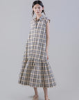 A young woman wears the Psophia Soft-Touch Plaid A-Line Drop-Waist Dress, crafted from a Tencel cotton blend with a ruffled hem, paired with beige ballet flats. Her long dark hair is styled in low pigtails against a plain light gray background.