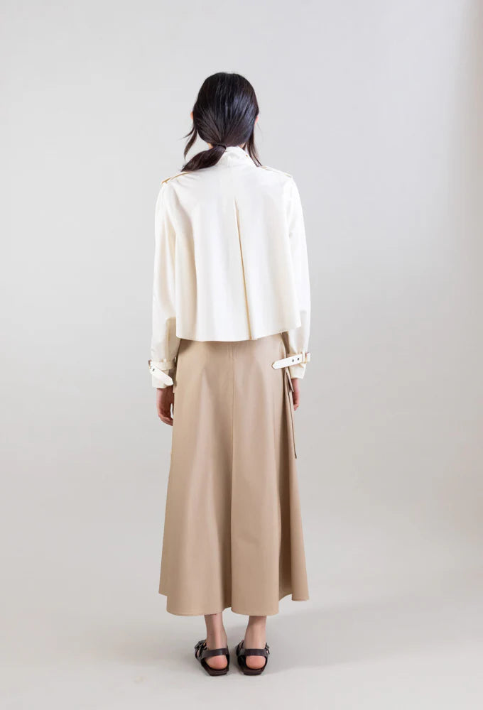 A woman with long dark hair stands facing away, wearing the Psophia Cropped Scarf Jacket. She pairs it with a beige A-line skirt and black sandals against a plain light gray background.