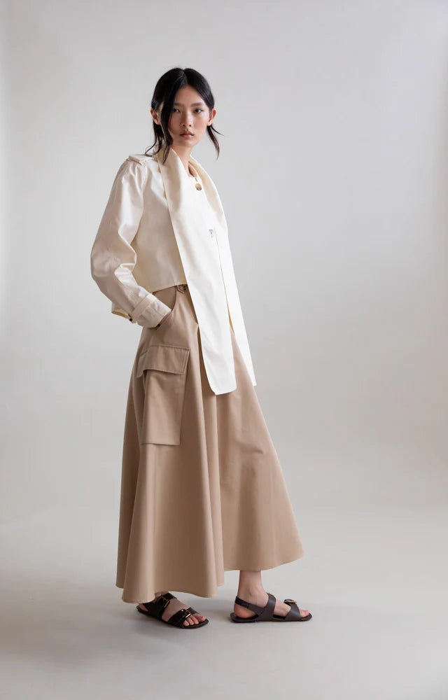 A woman stands against a plain background wearing the Psophia Cropped Scarf Jacket, paired with a beige, ankle-length skirt featuring large pockets. She has her hands in her skirt pockets and is wearing black sandals.