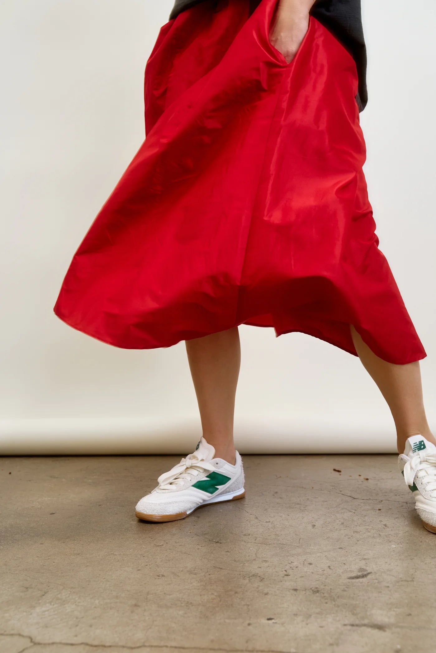 Wearing an Aquarius Cocktail LE SKIRT Silk Taffeta and white sneakers with green accents, a person stands on a concrete floor. Holding the skirt&#39;s edges, they create movement against an off-white wall.