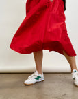 Wearing an Aquarius Cocktail LE SKIRT Silk Taffeta and white sneakers with green accents, a person stands on a concrete floor. Holding the skirt's edges, they create movement against an off-white wall.