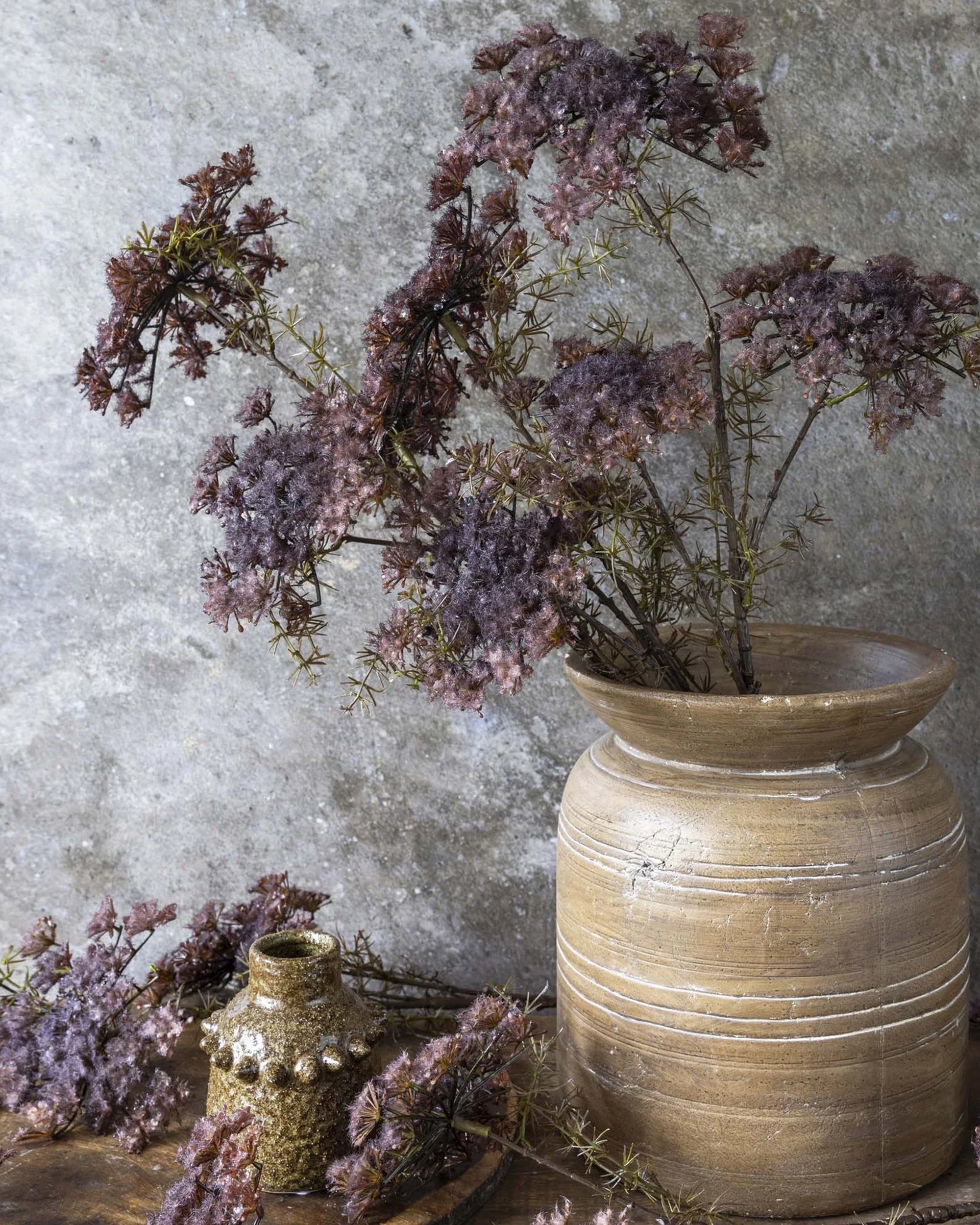 The Abigail Ahern Queen Anne's Lace Purple is arranged in a rustic ceramic vase against a textured gray backdrop, with a small ornamental gold jar and loose flower sprigs on the wooden surface nearby.